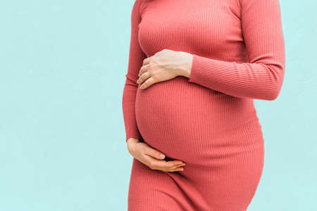 Pregnant woman in tight orange dress holds hands on belly isolated on blue background. Pregnancy and maternity concept. Close-up, copy spaceの写真素材