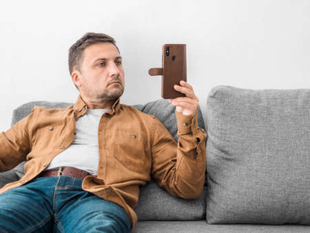 Middle aged man in beige shirt and jeans sits at home on gray sofa and looks intently at the phone....