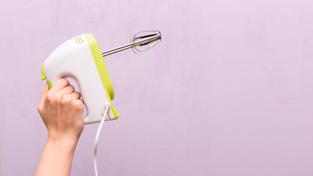Female hand is holding a mixer. Kitchen electric appliance on a purple background. Woman housewife. Empty space for textの写真素材