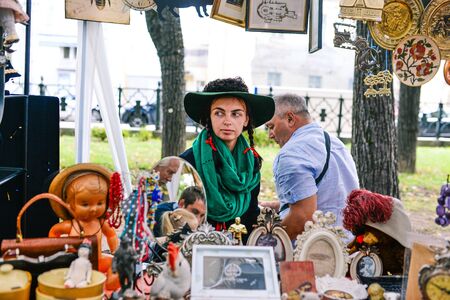Russia, city Moscow - September 6, 2014: Young beautiful girl in a hat with paly and green scarf. A woman sells antiques in a street market. Girl seller at a flea marketのeditorial素材