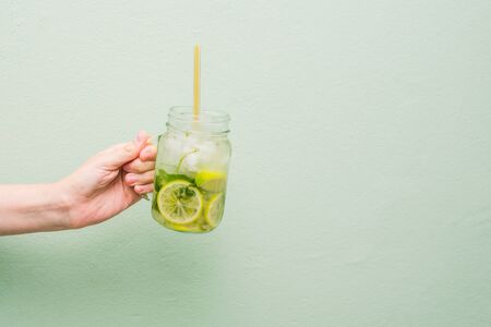 The hand of a young girl holds out a cocktail. Summer fruit drink in a glass jar with lime, rum and soda. Cold alcoholic mojito. The concept of relaxation, party and good mood. Copy spaceの写真素材