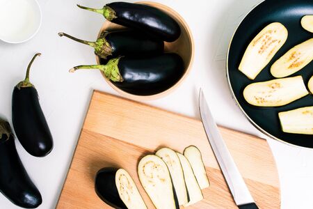 Cooking fried eggplant in a pan. Sliced and whole eggplant on a white background. Wooden board and knifeの写真素材