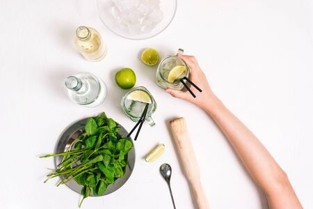 Girl holds a fresh mojito cocktail in her hand. Ingredients for making a summer drink on a white table. Top view, flat layの写真素材