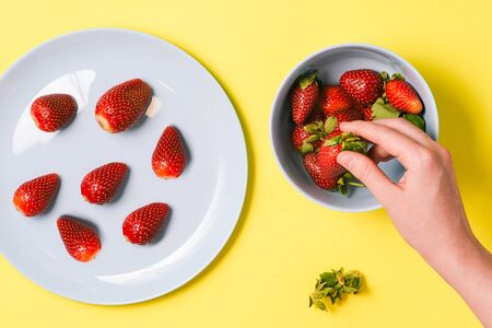 Girl's hand holds strawberries on a yellow background. Blue dishes with strawberries. Healthy summer meals for breakfast, lunch or dinner. Top view, flat layの写真素材