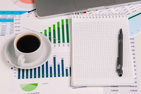 Cup of coffee, a computer, a notepad with a pen, and statistical graphs on the desktop. Analysis and monitoring of data, profit growth and cost planning of the companyの写真素材