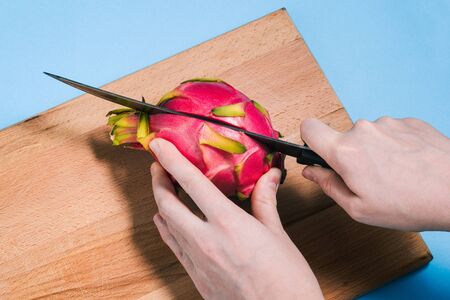 Close-up of a womans hands cutting a ripe pitaya fruit with a knife on a wooden Board. Pattaya is cut into two parts. The view from the top.の写真素材