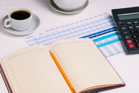 Open diary with a blank page and a pencil. Desktop with a calculator, a table with numbers and a Cup of coffee. Financial document. Monthly or annual report of the company.の写真素材
