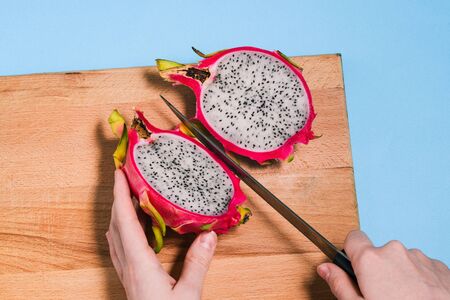 Pitaya cut into two halves. Close-up of female hands cutting summer dragon fruit. Pitaya on a wooden Board with a blue background.の写真素材