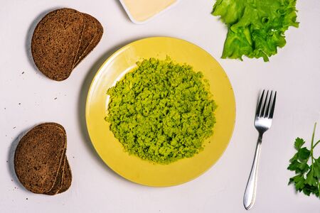 Making avocado toast. A white table with a yellow plate and mashed avocado, dark bread, a fork, and lettuce and parsley leaves. Recipe for avocado sandwiches. A top view of a flat layout.の写真素材