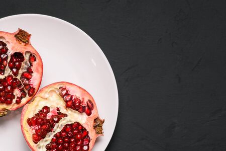 Cut into two parts ripe pomegranate on a white plate. Red garnet with grains on a black background with texture. Top view, copy space.の写真素材