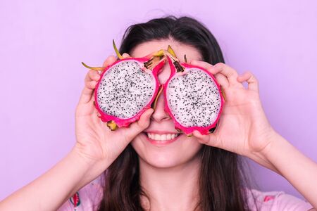 Young girl holds a pitaya near her eyes and smiles. Smiling woman with fruit on a purple background. Ripe tropical dragon fruit.の写真素材