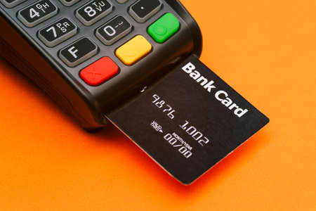 Close-up of a payment terminal with a credit card. Modern device for paying for services and goods on an orange background. Writing off money from a debit cardの写真素材