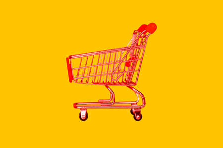 Shopping trolley in the store. Red basket on a yellow background. The concept of retail, sale of goods and products. Food trolley. Food delivery by courier from the supermarketの写真素材