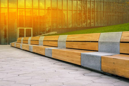 Wooden bench and tiled walkway. Office building for the work of company employees. Glass walls of a building with construction site reflection. Interior elements. Modern urban landscape.の写真素材