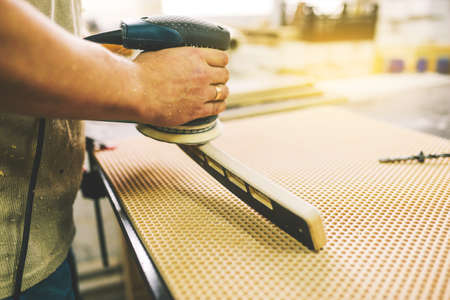 A man processes a wooden part with a grinder. Manufacturing of furniture in a carpentry workshop. Wood polishing machine. Hobby, manual labor and freelance concept.の写真素材