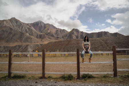 Brunette European girl sits on a ranch against a background of mountain. Wild West. Autumn. Wideの写真素材