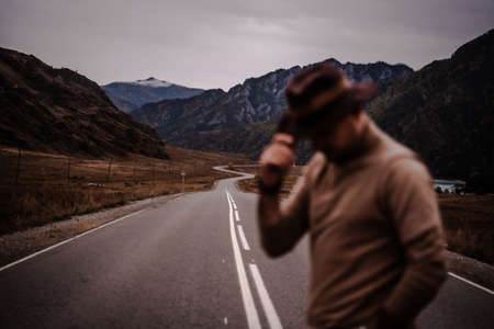 Young stylish guy in a hat stands on the background of a mountain road. Autumnの写真素材