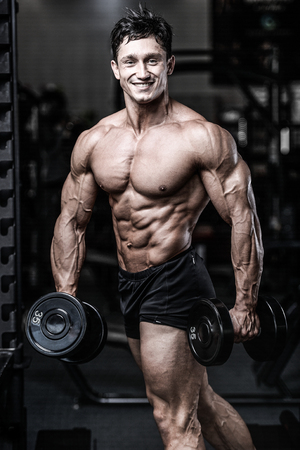 Brutal bodybuilder on diet resting and working out in the gym. Caucasian sexy fitness male model execute exercise with dumbbellsの写真素材