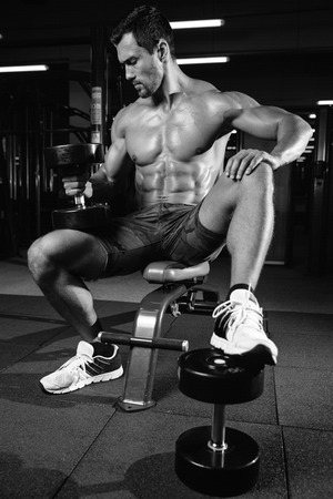 Brutal bodybuilder on diet resting and working out in the gym. Caucasian sexy fitness male model execute exercise with dumbbellsの写真素材