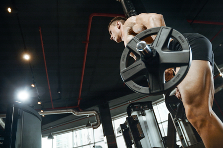 Handsome young muscular Caucasian man of model appearance working out training pumping up back lats muscles in the gym gaining weight on machines posing fitness and bodybuilding conceptの写真素材