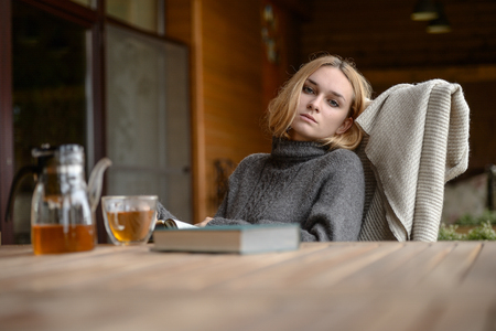 cute well dressed pretty young caucasian blonde hair adult teenage girl in fashion dress at farm house with cup of tea and book at countryside landscape spring or autumn weather depressed moodの写真素材