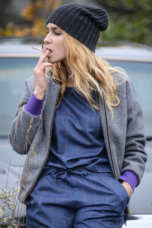 cute well dressed pretty young caucasian blonde hair adult teenage girl in fashion dress near car smoking cigarette at countryside landscape spring or autumn weather depressed moodの写真素材