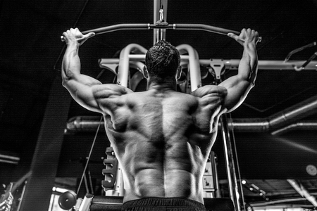 Handsome young muscular Caucasian man of model appearance working out training pumping up back lats muscles in the gym gaining weight on machines posing fitness and bodybuilding conceptの写真素材