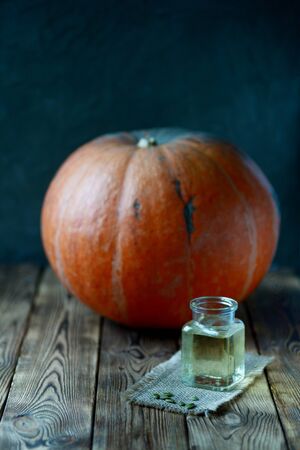 Yellow pumpkin oil in glass transparent opened jar in front of big orange pumpkin raw vegetable seeds and bottle on wooden brown table dark background autumn still life healthy organic nutritious foodの写真素材