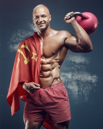Bodybuilder handsome strong athletic rough man pumping up muscles with kettlebell workout fitness and bodybuilding healthy concept soviet flag background - muscular man doing exercises in gymの写真素材