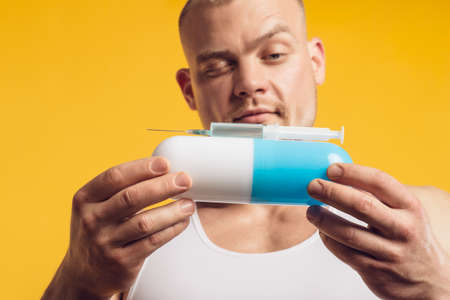 Studio portrait of muscular man with pill and syringe, injector, standing over isolated yellow background. Fitness, bodybuilding and work out conceptの写真素材