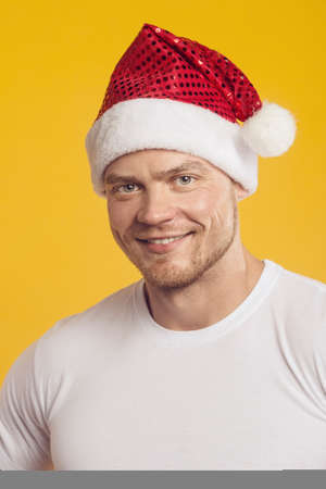 Studio portrait of muscular man in santa claus hat and present box in white shirt, standing over isolated yellow background. Fitness, bodybuilding and work out conceptの写真素材