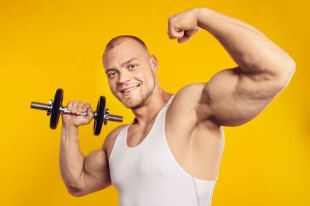 Studio portrait of muscular man with dumbbell in white shirt, standing over isolated yellow...