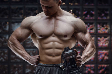 Fitness man in gym drinking water after workout looking at cell phone. Fitness and bodybuilding healthy background. Caucasian men doing exercises in gym. Water bottle and supplements nutrition conceptの写真素材