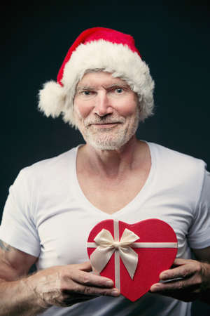 Portrait of senior man in Santa Clause hat in Gym holding a gift in his hands. Elderly age sport and fitness concept. New Years eve and Christmas backgroundの写真素材