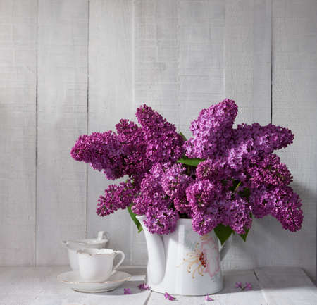 Cup of coffee and Lilac Bouquet in ceramic jug against a white wooden board.の写真素材