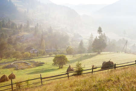 Rural landscape in foggy dawn with wooden fence and haystacks on the meadow in the foreground,...