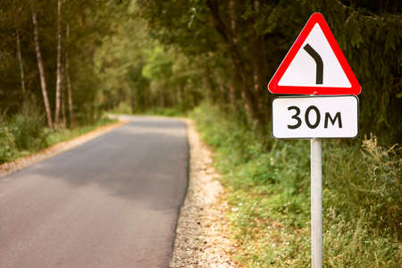 Road sign dangerous turn after 30 meters on a car road in the forest with a blurred backgroundの写真素材