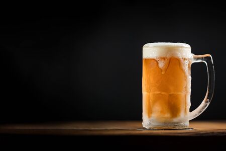 cold mug with beer, with overflowing foam, on wooden table and dark background wish spaceの写真素材