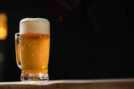 cold mug with beer, with overflowing foam, on wooden table and dark background wish spaceの写真素材