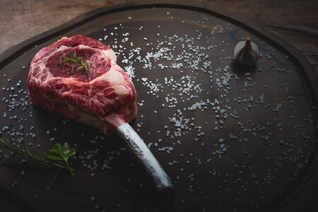 raw tomahawk beef steak with ingredients for grilling with a spice rub, fresh rosemary and salt , on a table with dark wood textureの写真素材