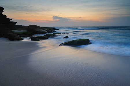 Sunrise seascape with a lone mossy rock with a flowing wave around itの写真素材