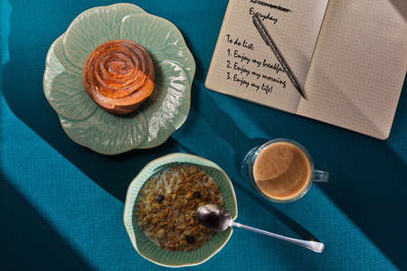 Light luncheon consists of muesli, cinnamon bun and coffee shooted on an aquamarin table near the notepad with some kind of a planning for day offの写真素材