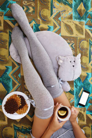 Girl sitting in bedroom in knee-height socks with a cup of tea, waffels, toy cat and a cell phone