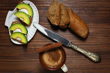 Sandwiches with spreading cheese and avocado slices on shell like plate with antique knife and coffee with cinnamon on the oldened wooden tableの写真素材