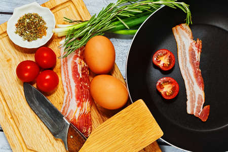 Rural style concept of omelette or fried eggs preparation with pan, different ingredients including pork bacon, cherry tomatoes, herbs and spices mixの写真素材