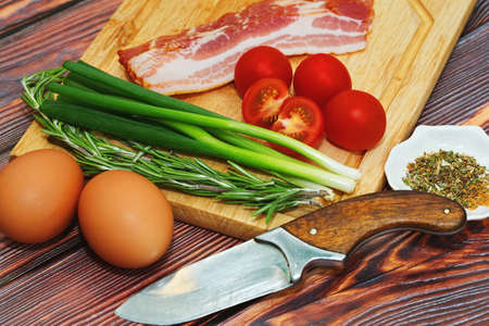Some ingredients for nutritious meal cooking - eggs, pork bacon, tomatoes, green onion and rosemary - close up caption in simple rural styleの写真素材
