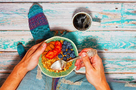 Person in ripped jeans and cozy socks is sitting on wooden floor and having his healthy dieting...