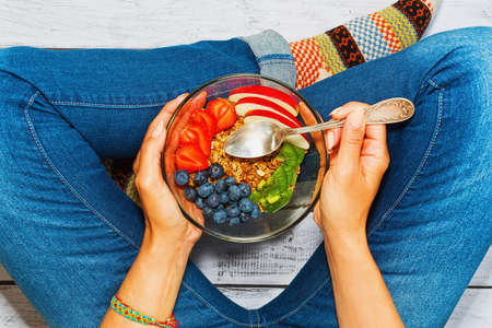Young female person sitting cross legs on wooden surface with a bowl of nutritious breakfast - granola with apple, kiwi, blueberry, strawberry, almonds. Healthy lifestyle concept. First person view.の写真素材