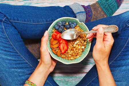 Young person having healthy breakfast of granola with berries and nuts while sitting cross-legged on the wooden floor in cute warm socks. Cosiness and healthy lifestyle concept. First person view.の写真素材