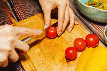 Young female person slicing tiny cherry tomatoes for salad on cutting board on wooden kitchen table....